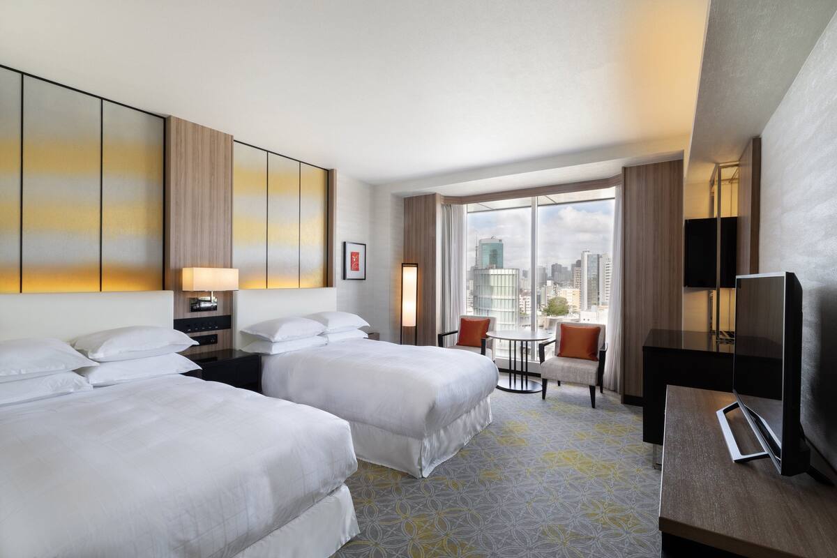 A hotel room with two beds, a desk, a chair, and a TV.