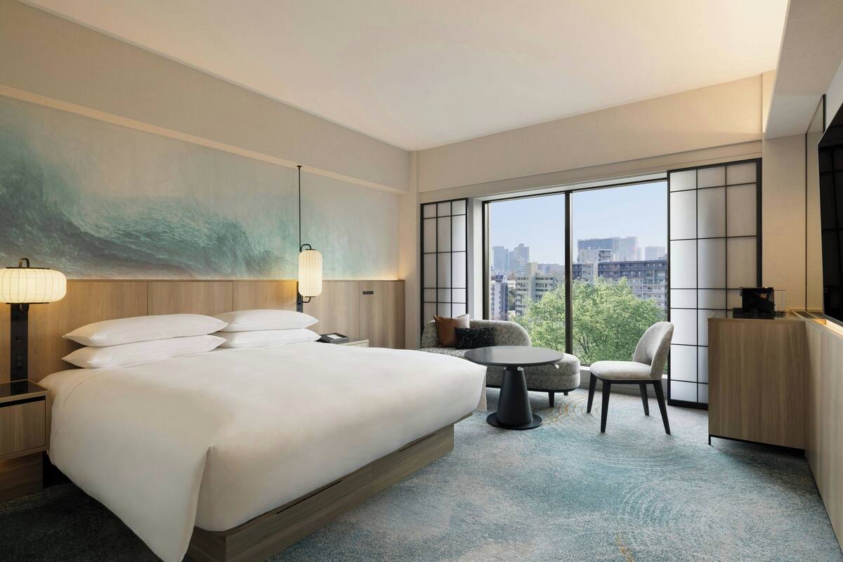 A hotel room with a large bed, a desk, a chair, a TV, and a view of the city through large windows.