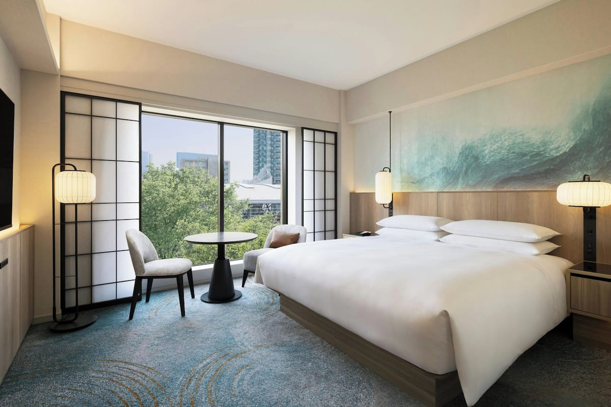 A hotel room with a large bed, two bedside tables, a chair, a small round table, and a large window offering a view of buildings and trees.