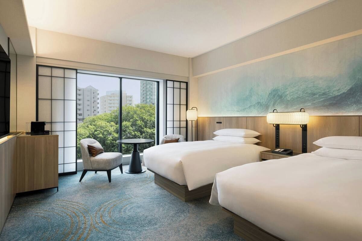 A hotel room with two beds, a desk, a chair, a small table, and a large window offering a view of buildings and trees.