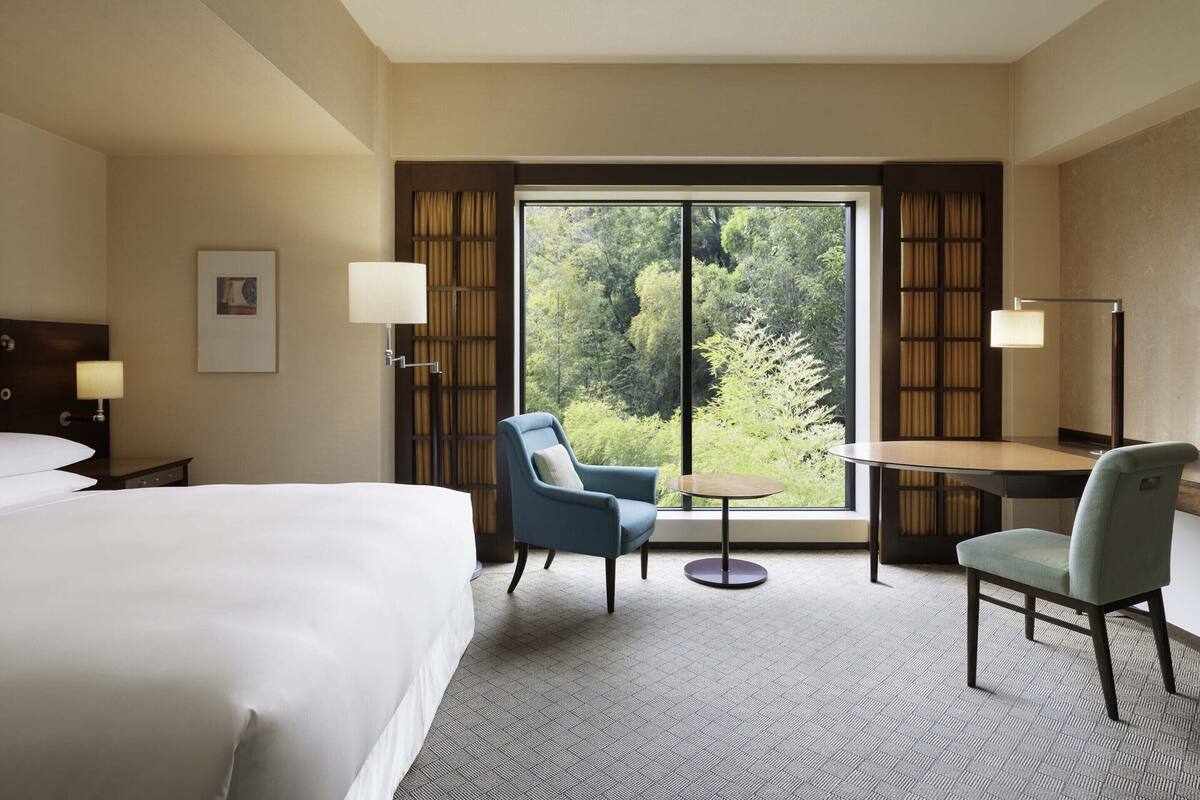 A hotel room with a large bed, two chairs, a small table, and a view of a forest through a large window.