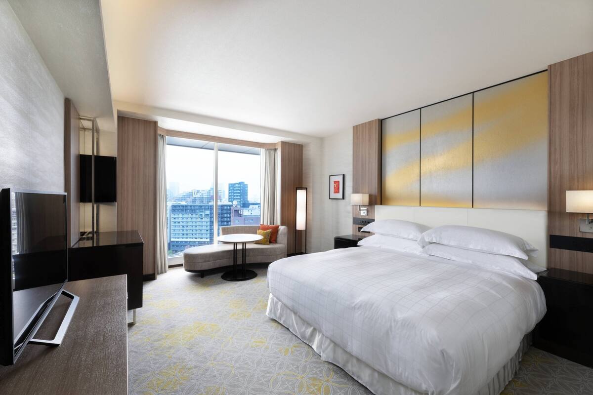 A hotel room with a large bed, a desk with a flat-screen TV, a small round table, and a city view through the window.