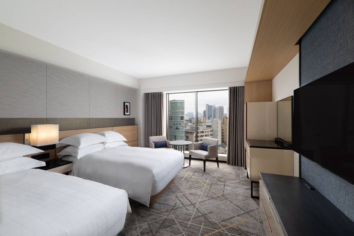 A hotel room with two beds, a TV, a small table, and a view of the city.
