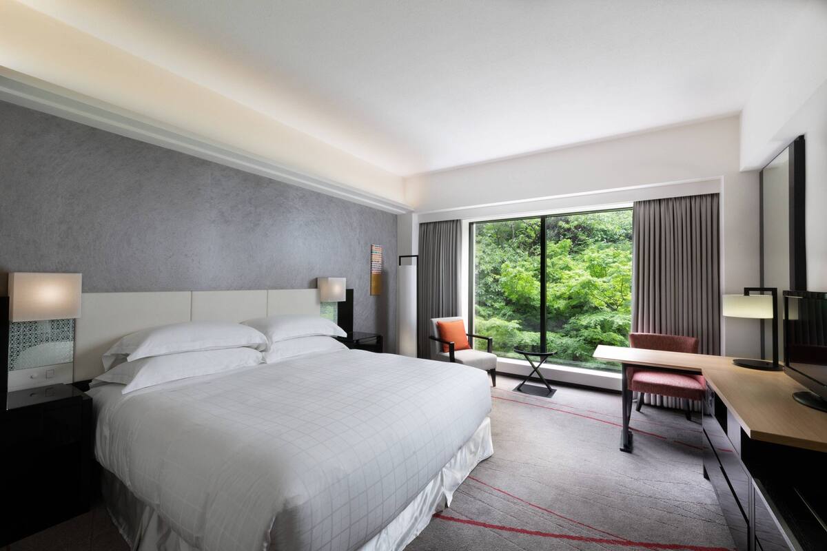 A modern hotel room with a large bed, a desk, and a view of greenery through the window.