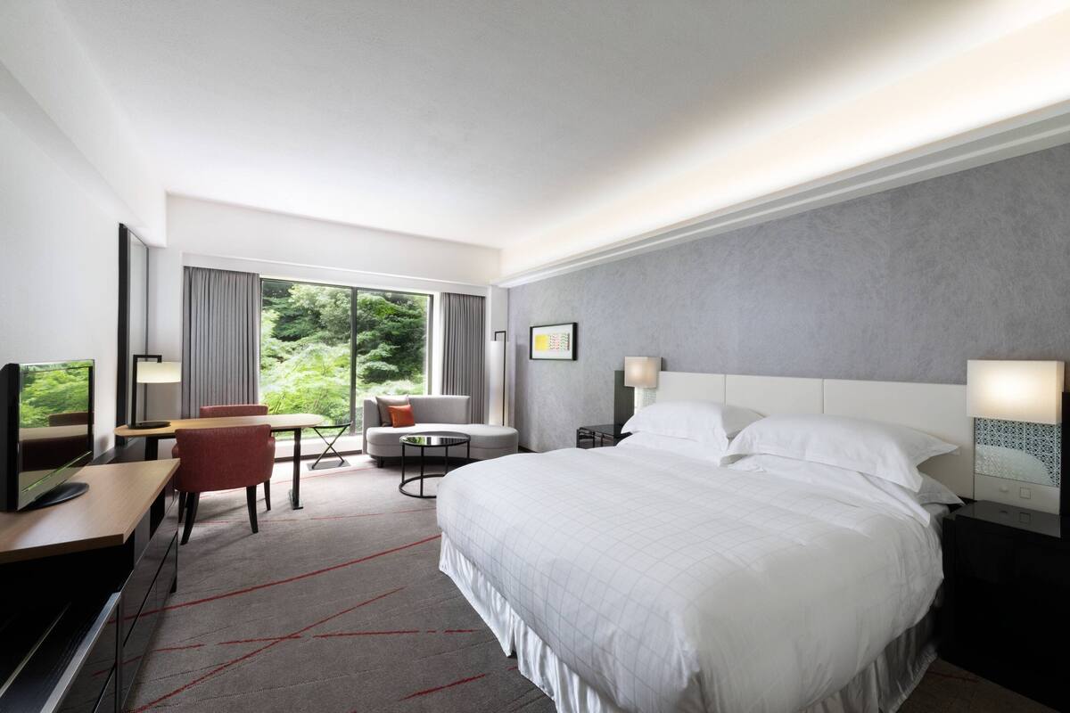 A modern hotel room with a large bed, a desk with a computer, a chair, and a sofa.