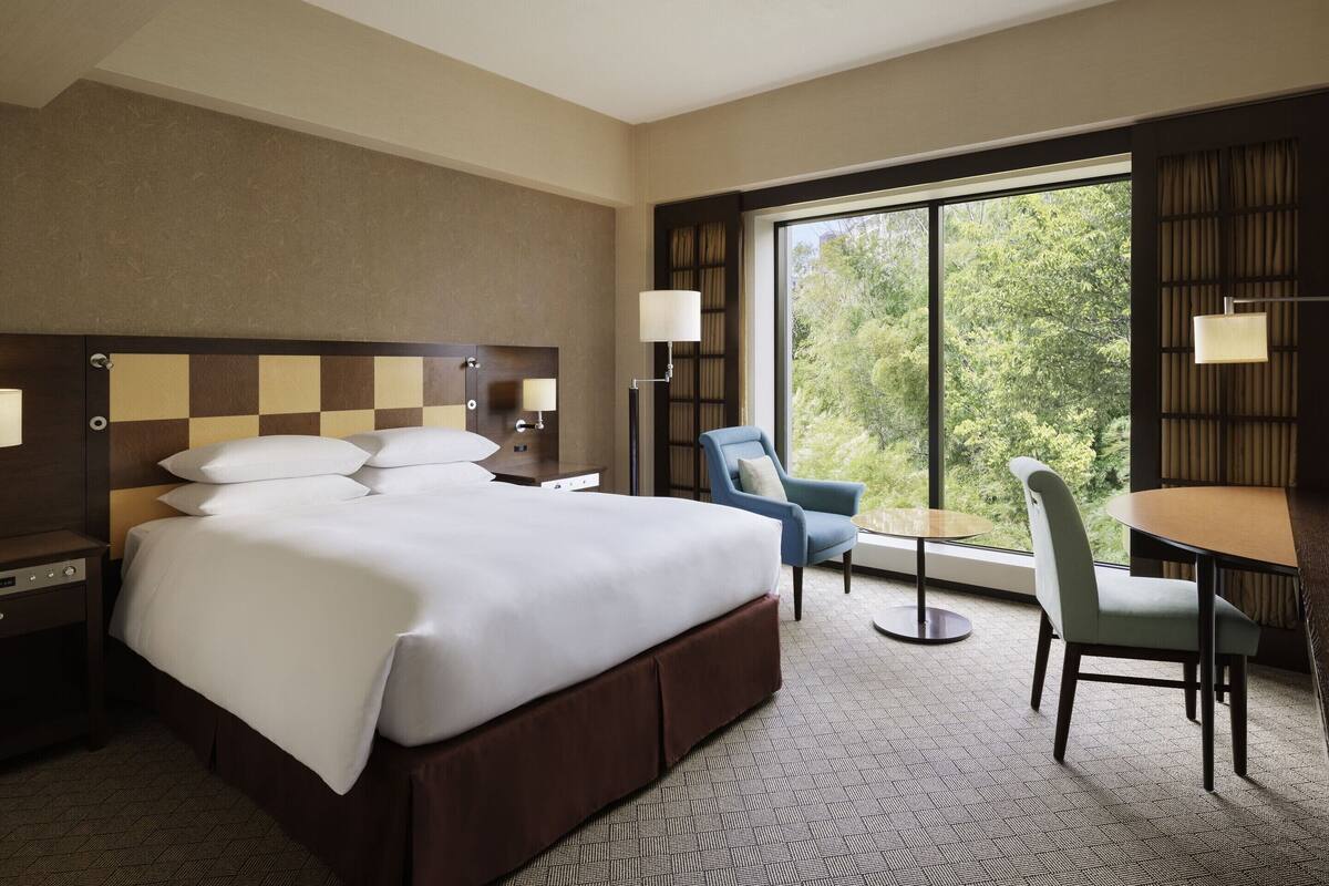 A hotel room with a large bed, a desk, a chair, and a view of trees through the window.