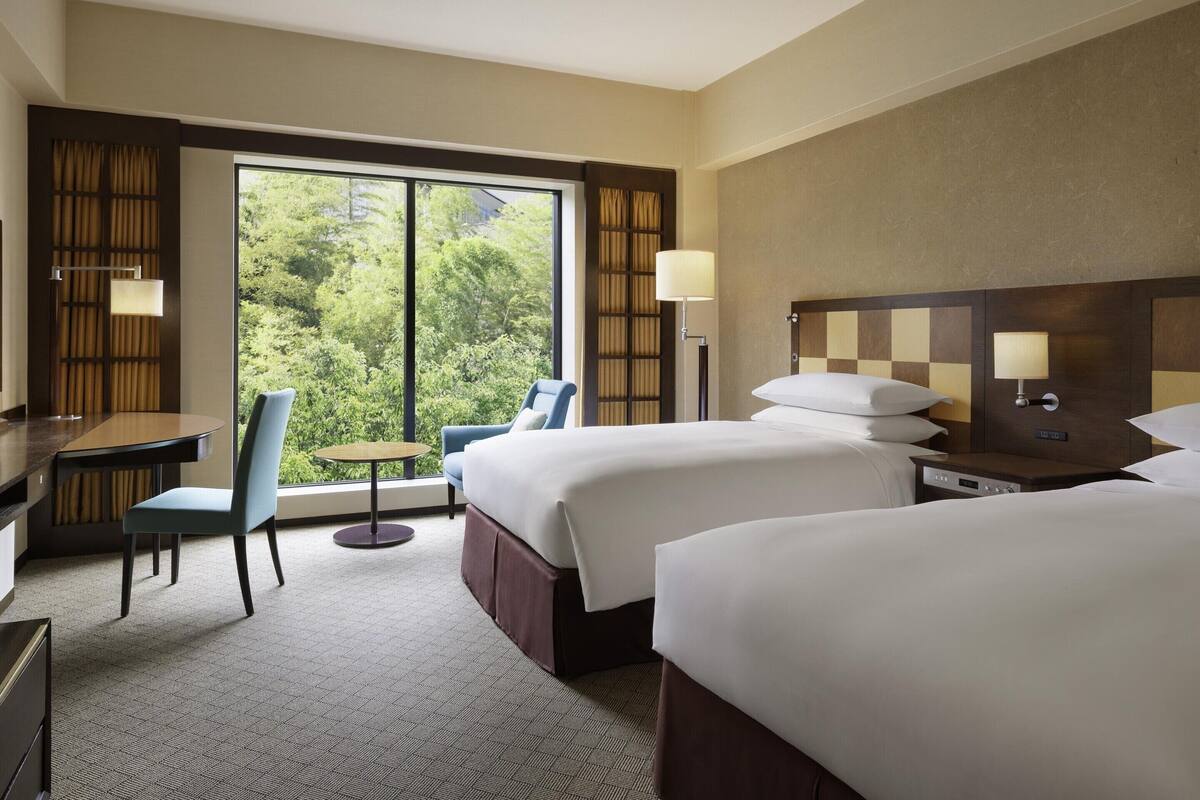 A hotel room with two beds, a desk, a chair, and a view of greenery through large windows.
