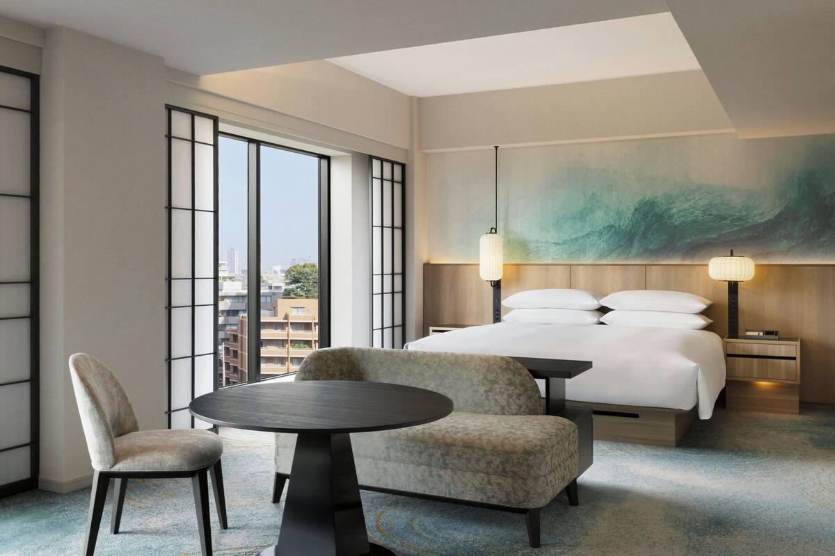 A hotel room with a large bed, a round table, two armchairs, a nightstand, and a large window offering a city view.