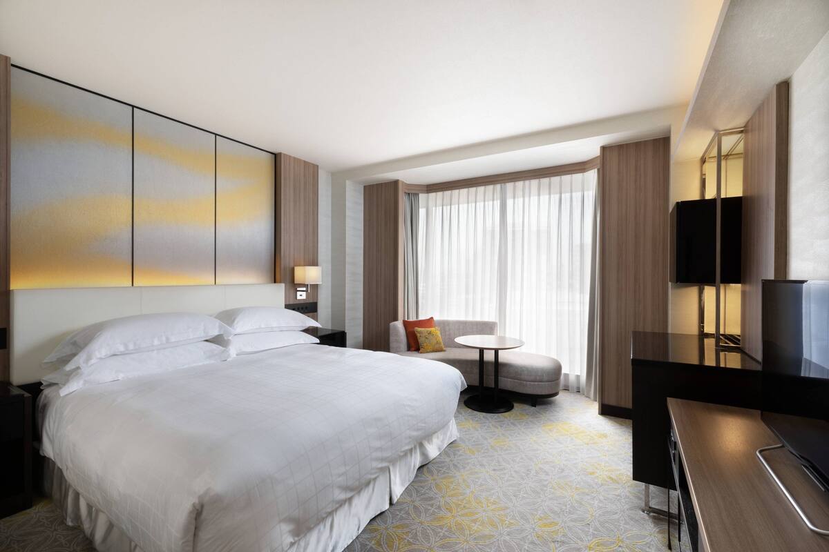 A modern hotel room with a large bed, a small round table, a sofa, a TV, and a large window with curtains.