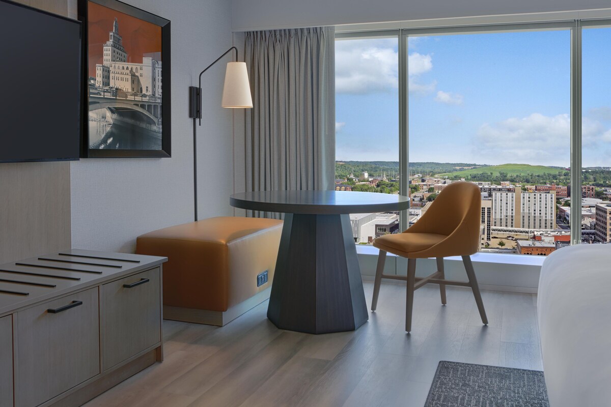 Habitación de hotel con escritorio, una silla, televisor montado en la pared y un ventanal con vistas a la ciudad.