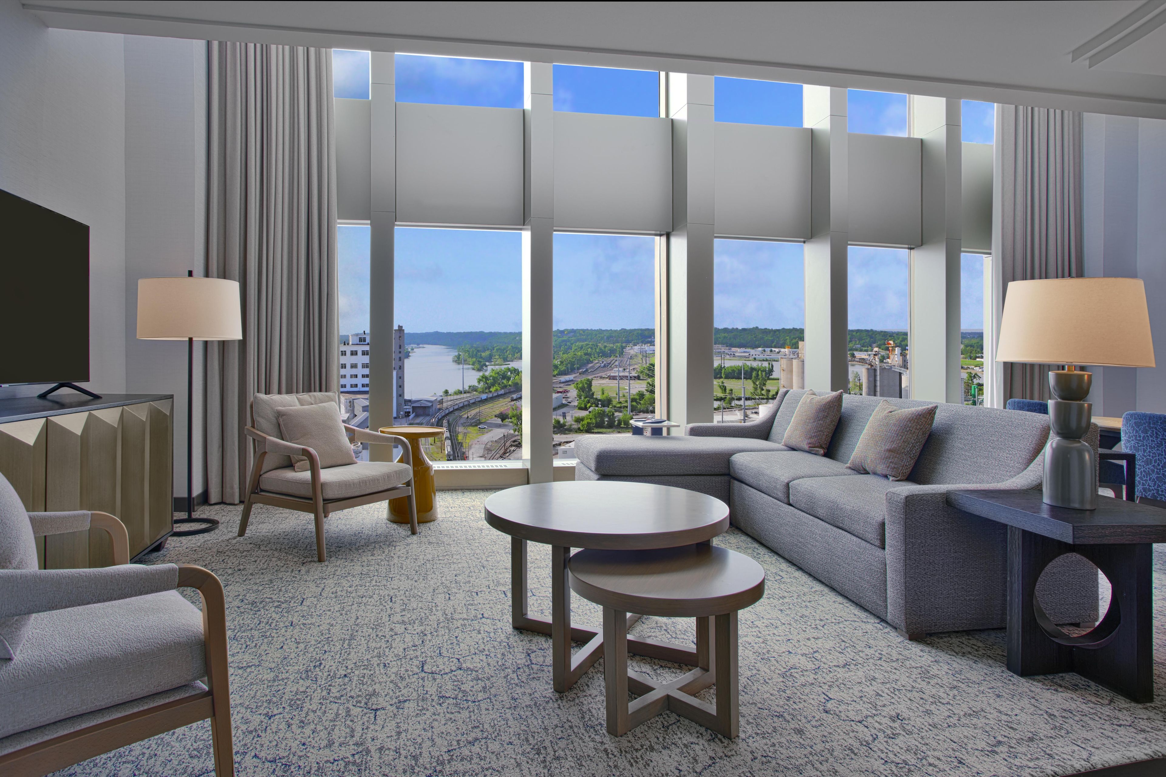 A modern hotel room with a large window offering a city view, a sofa, a chair, a small table, and a TV.