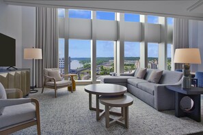 Habitación de hotel moderna con un gran ventanal que ofrece vistas a la ciudad, un sofá, una silla, una mesita y un televisor.