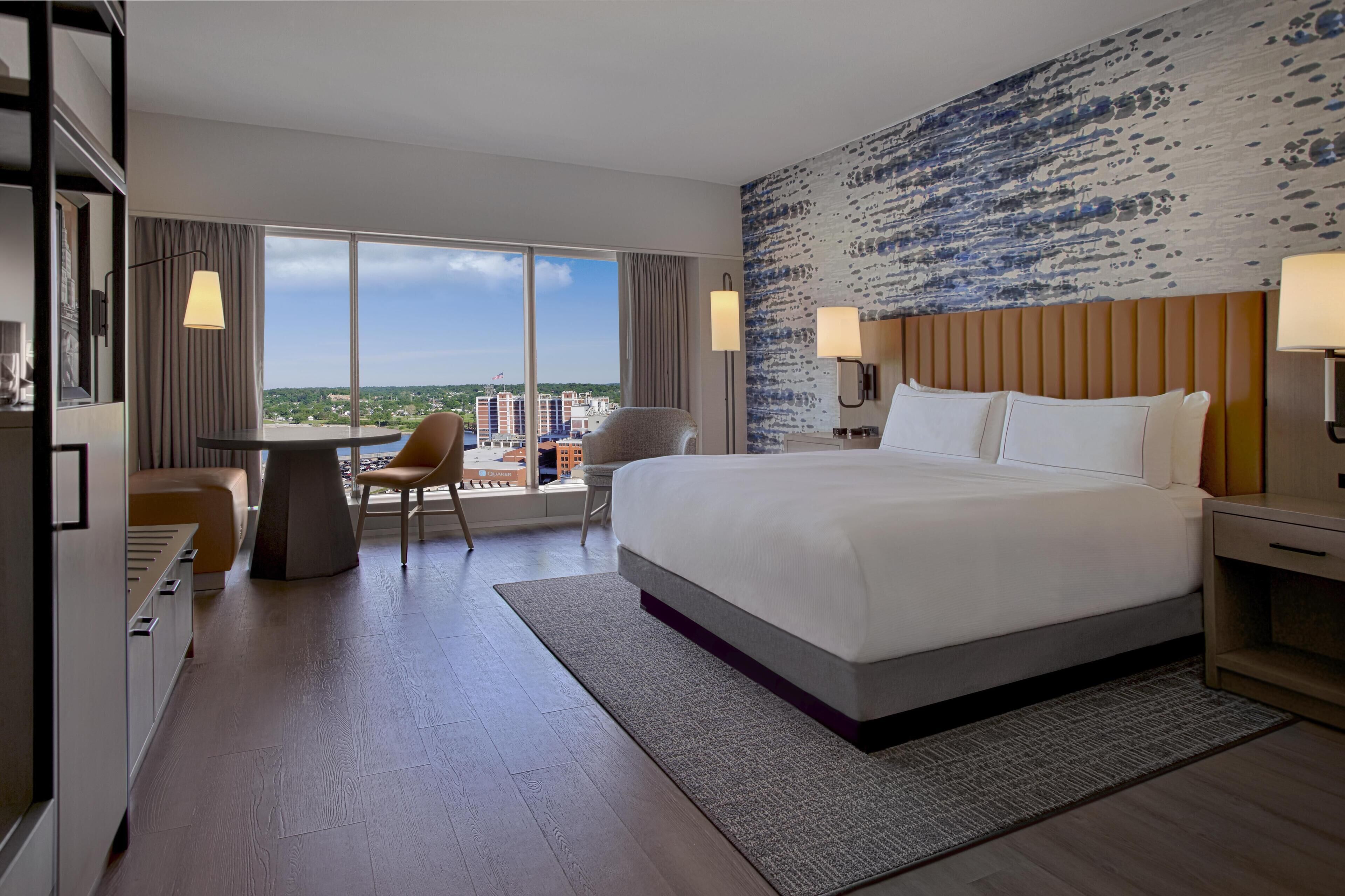 A hotel room with a large bed, a desk, a chair, and a view of the city.