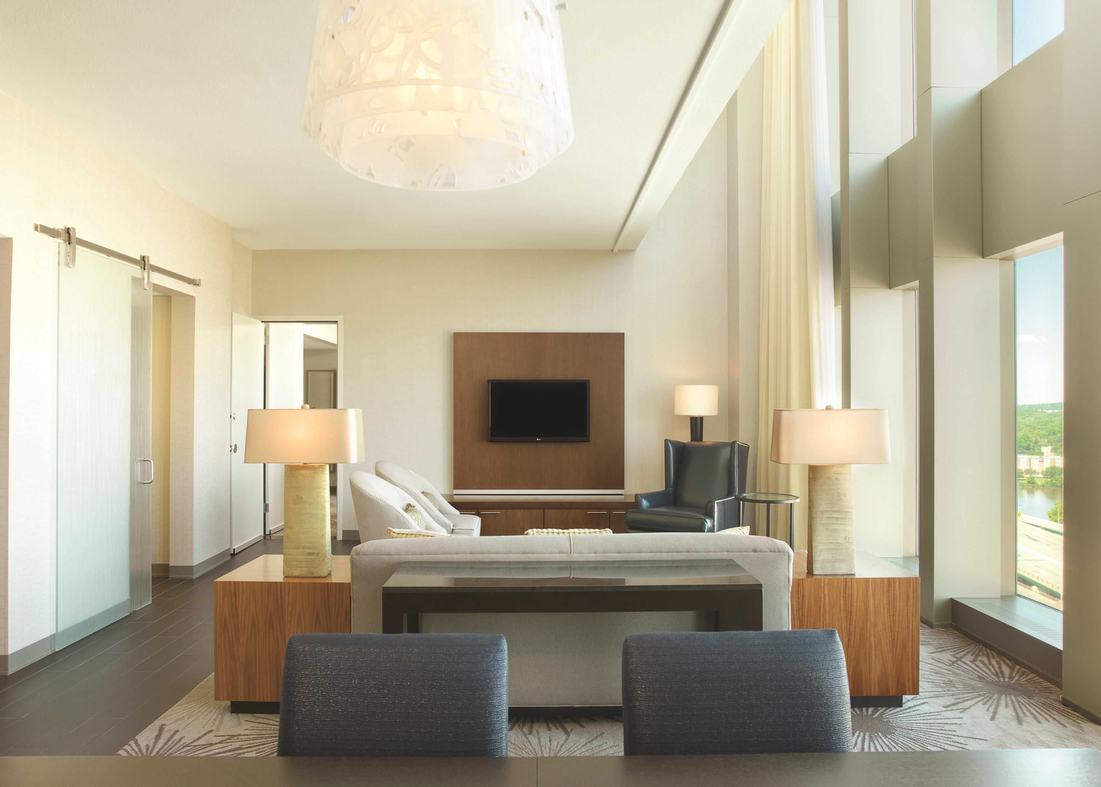 Une chambre d’hôtel moderne avec un grand lit, deux fauteuils, un téléviseur fixé au mur et une vue sur une rivière.