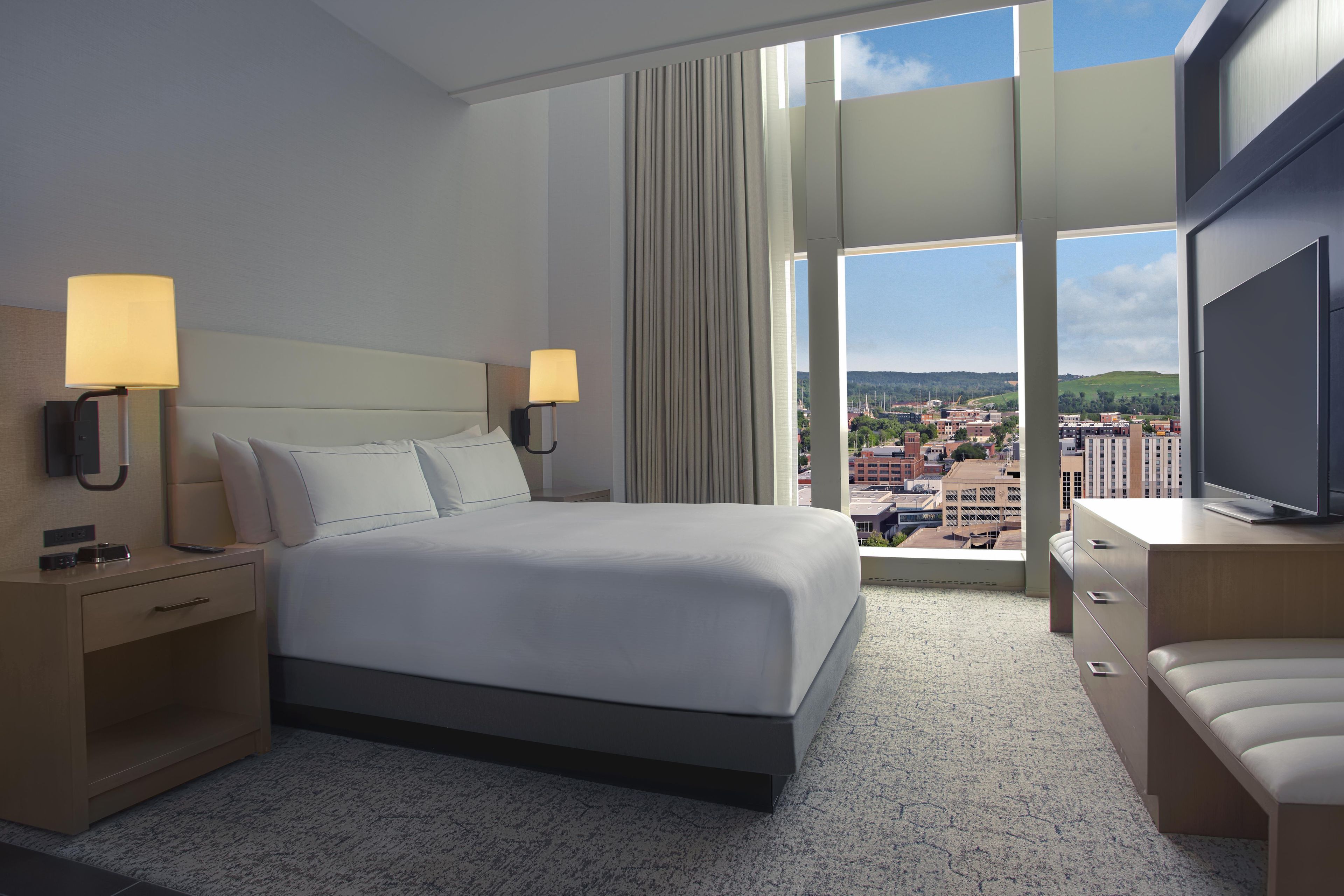 Habitación de hotel con una cama grande, dos lámparas de noche, un televisor y vistas a un paisaje urbano a través de amplias ventanas.