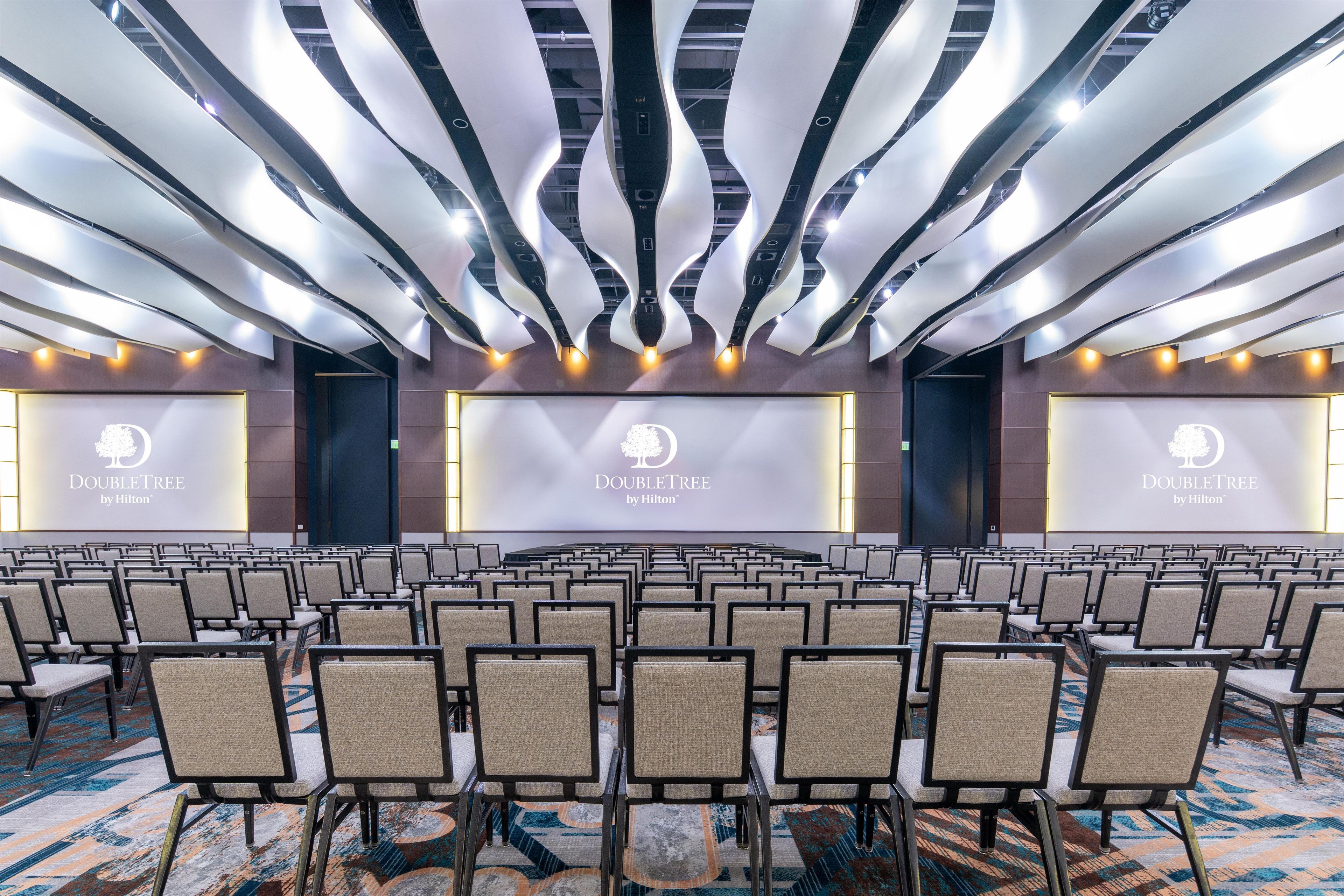 Ein Konferenzraum mit Reihen von Stühlen, einer Bühne mit einem Bildschirm, der das DoubleTree by Hilton Logo zeigt, und einem modernen Deckenstil.