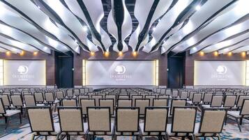 Ein Konferenzraum mit Reihen von Stühlen, einer Bühne mit einem Bildschirm, der das DoubleTree by Hilton Logo zeigt, und einem modernen Deckenstil.