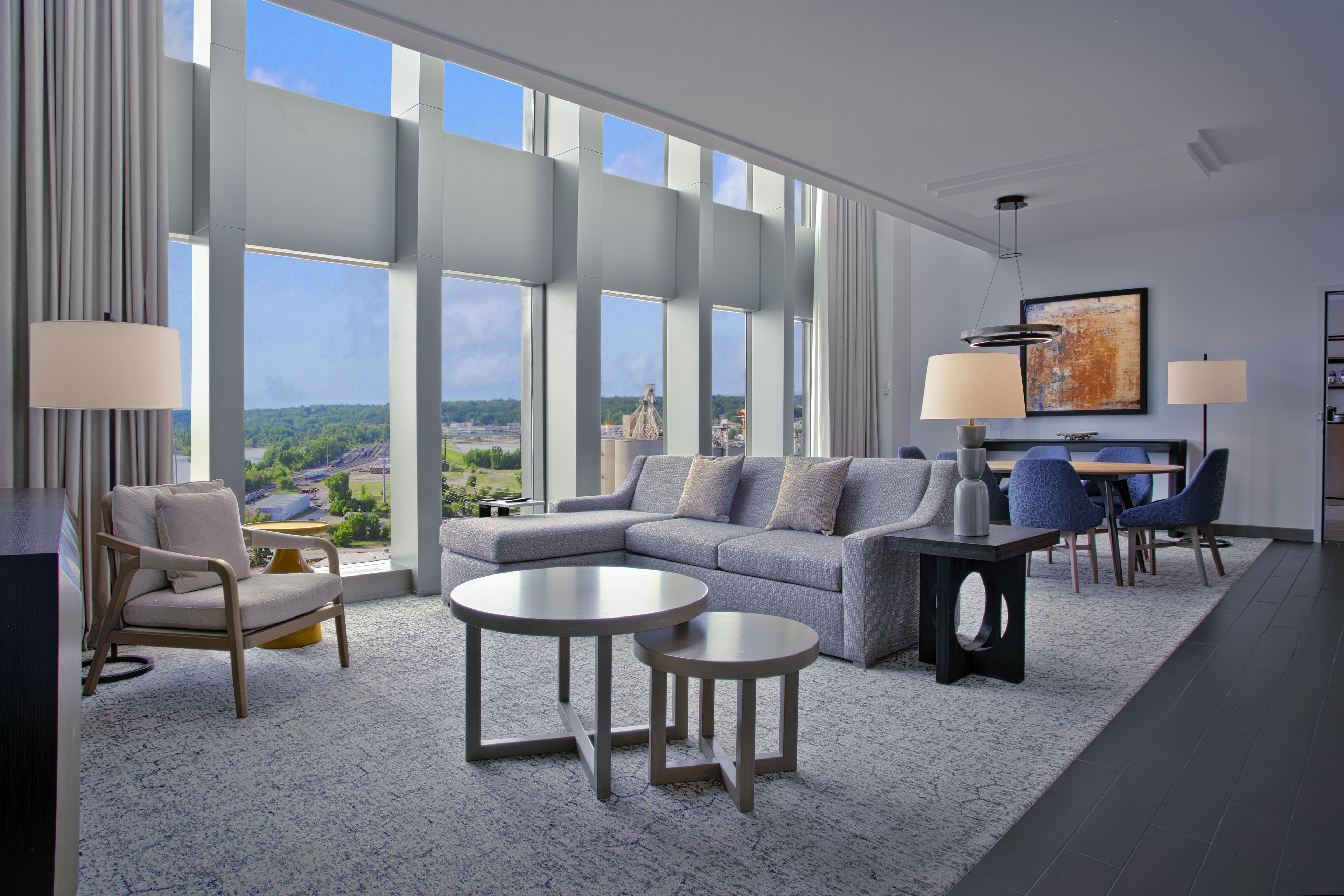 Uma sala de estar moderna com um sofá, duas mesas laterais, uma poltrona e um espaço de refeições com cadeiras. Janelas amplas oferecem vista para o exterior.