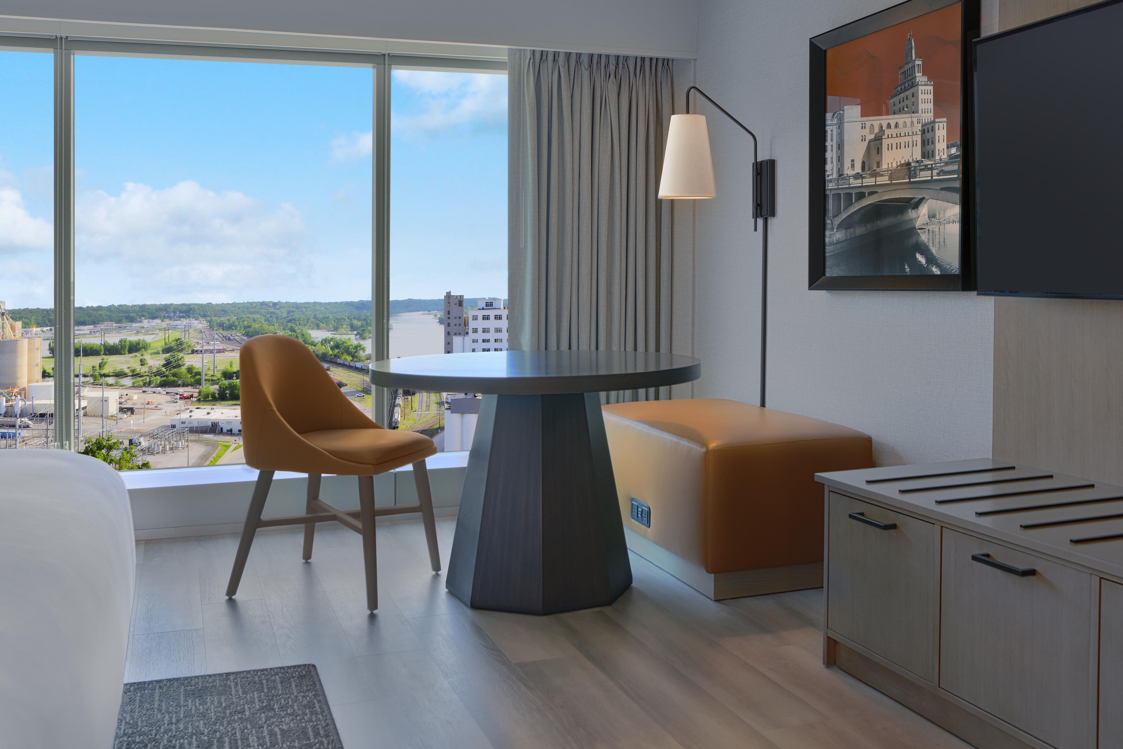 Habitación de hotel con un ventanal grande, un escritorio, una silla, un sofá, un televisor y un armario.