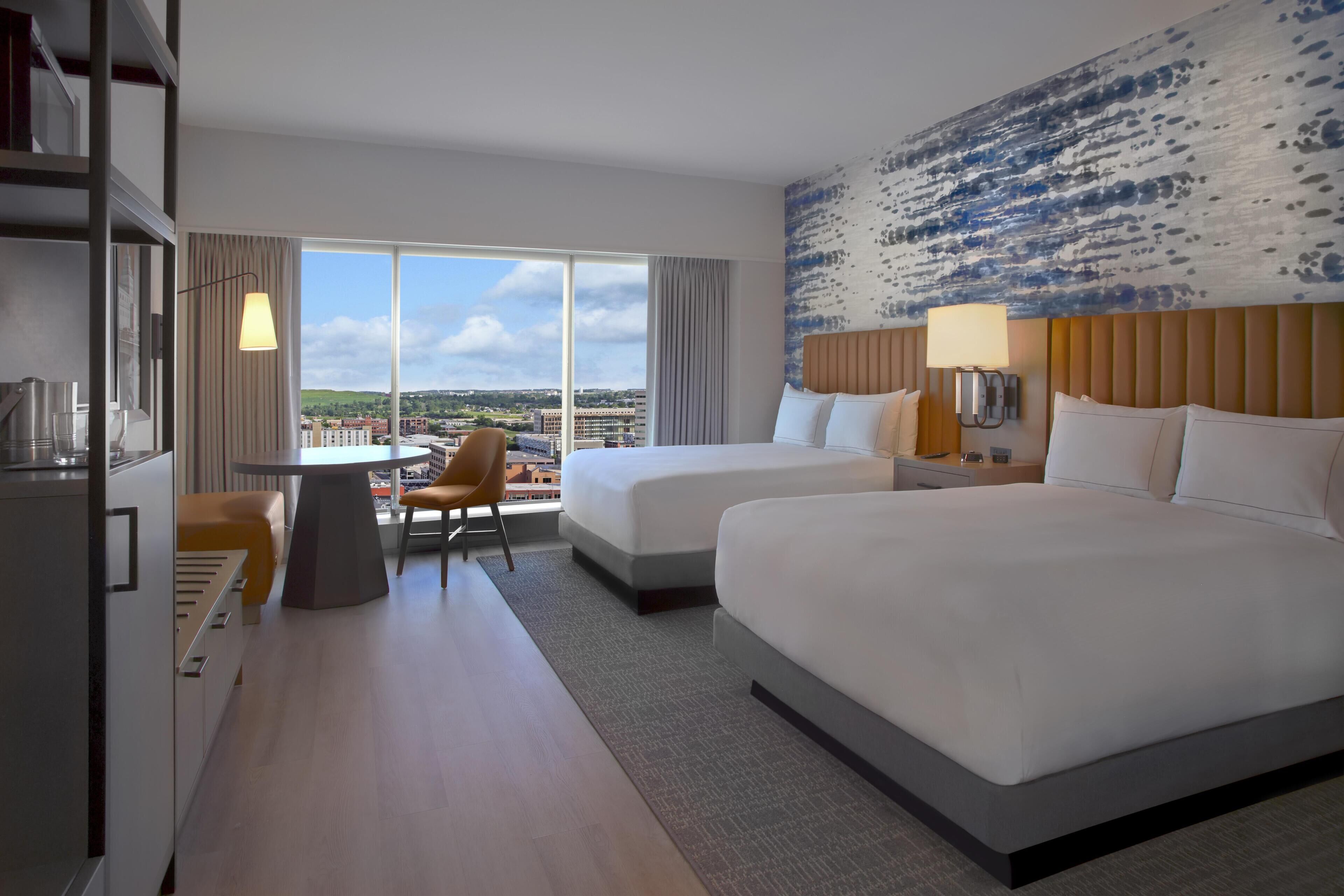 A hotel room with two beds, a dining table, a chair, and a view of the city.