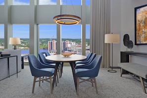Una habitación de hotel moderna con un ventanal grande, una mesa de comedor redonda, sillas azules, una lámpara de pie y un cuadro en la pared.