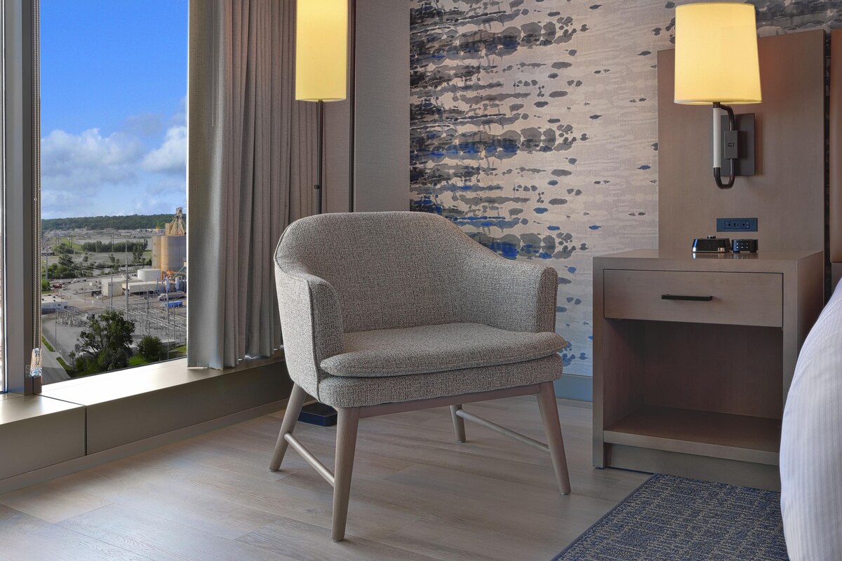 Habitación de hotel con vistas a un paisaje lejano, una silla moderna, una mesita de noche con una lámpara y una pared con textura.