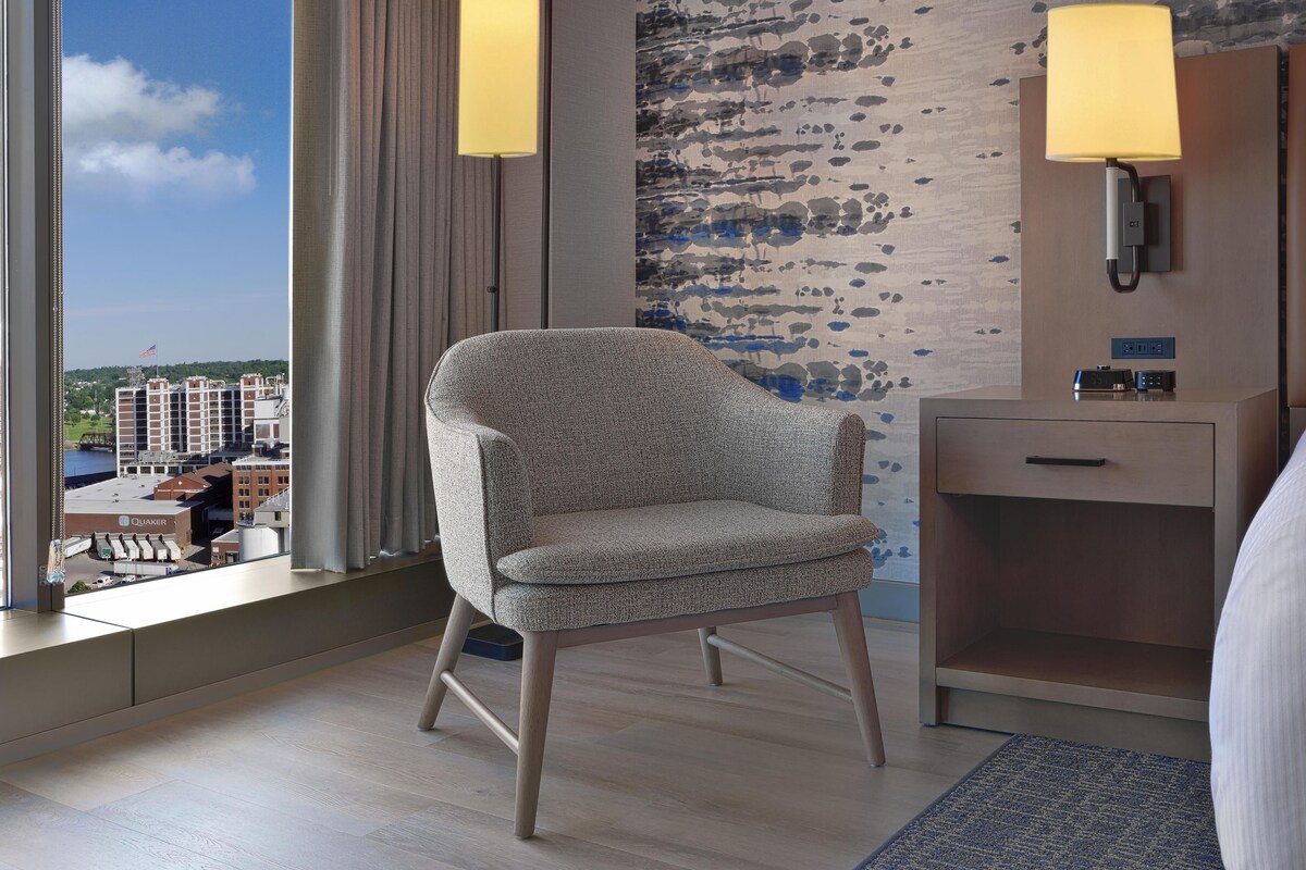 Habitación de hotel con vista a un paisaje urbano, un sillón estampado, una mesita de noche con lámpara y un teléfono en la pared.