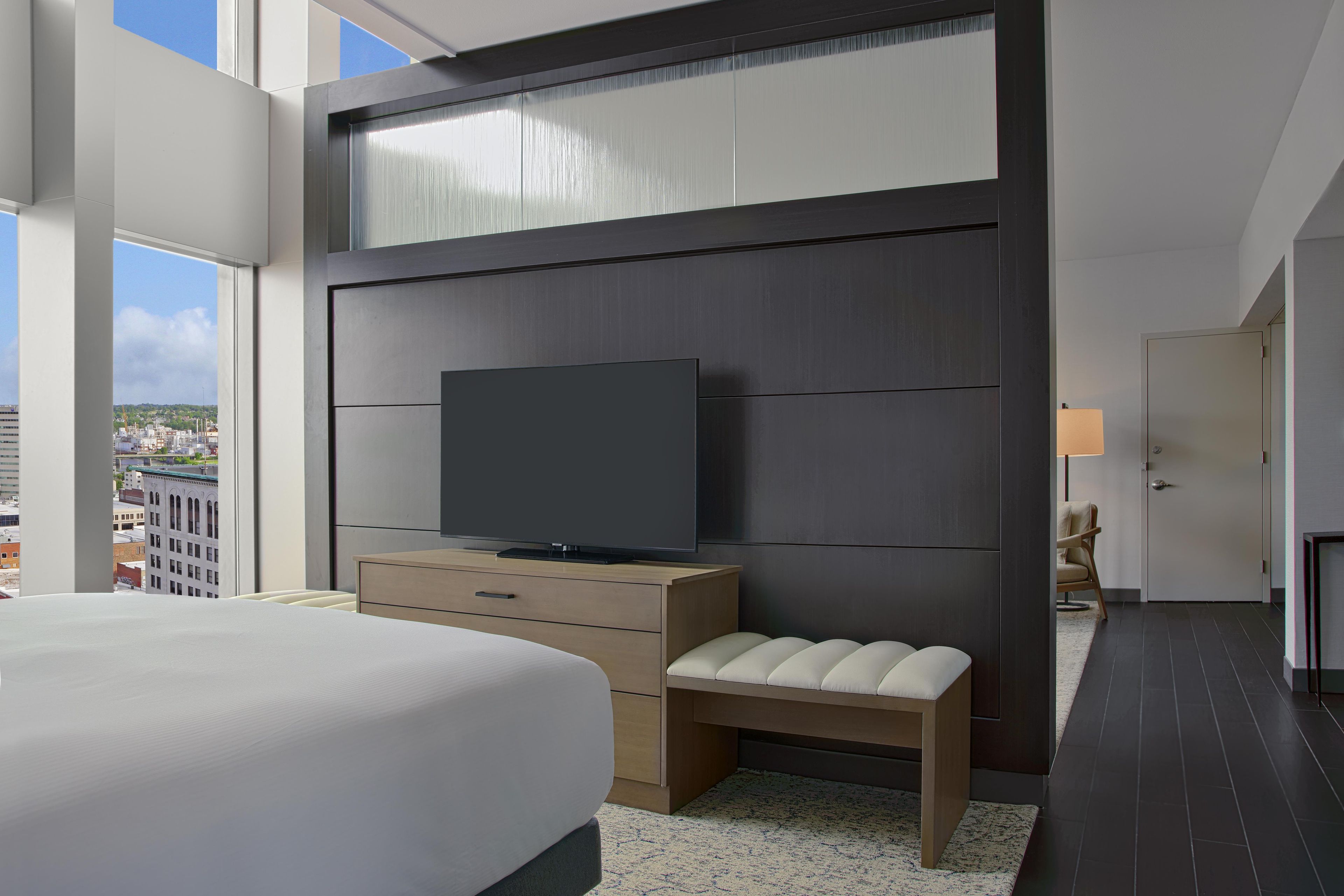 A modern hotel room with a large bed, a flat-screen TV, a bench, and a city view.