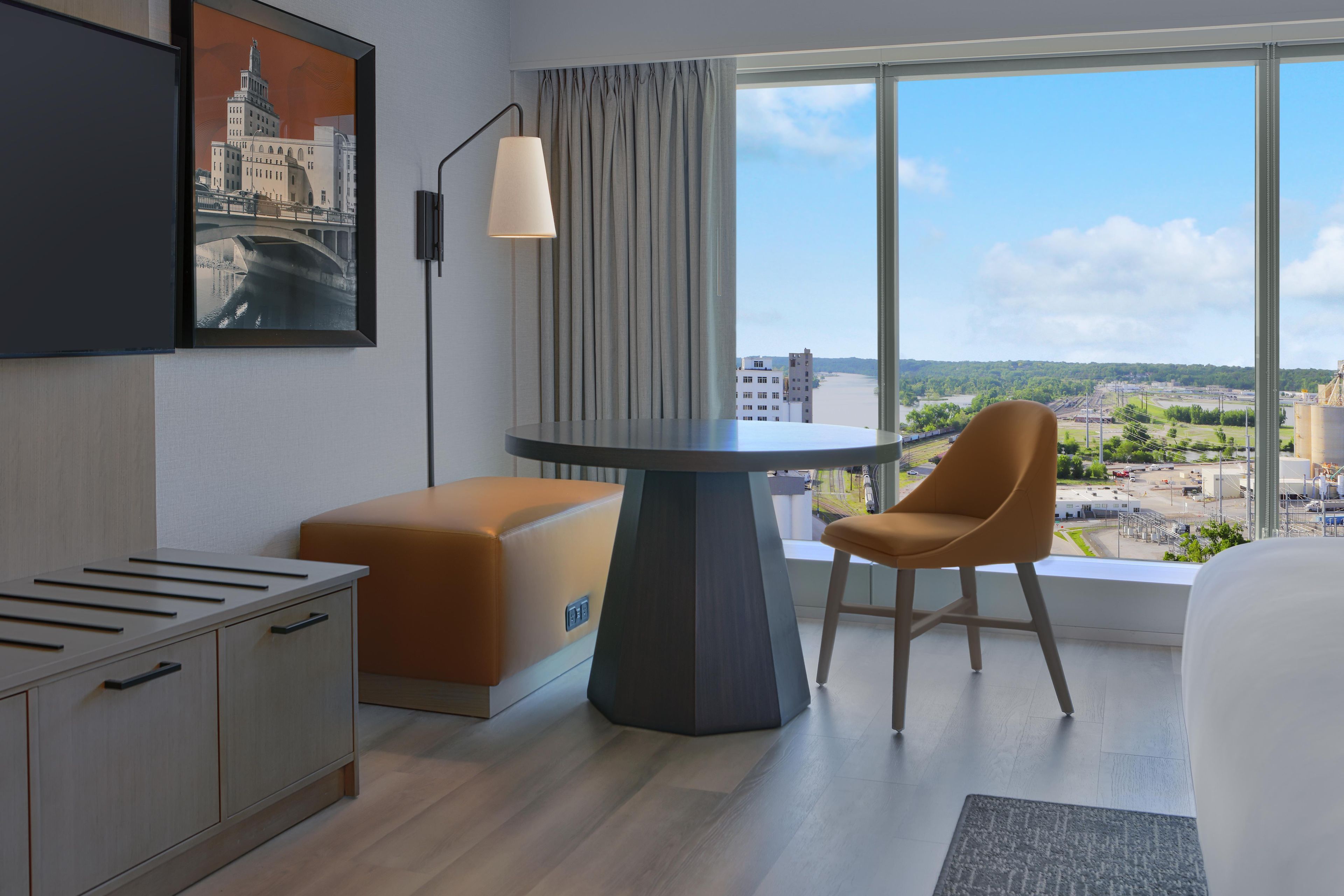 Een moderne hotelkamer met een bureau, een stoel, een bed, een aan de muur bevestigde televisie en een groot raam met uitzicht op de skyline.
