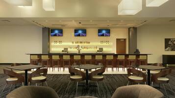 A hotel lobby with a reception desk, bar area, seating arrangement, and a television screen displaying a sports channel.