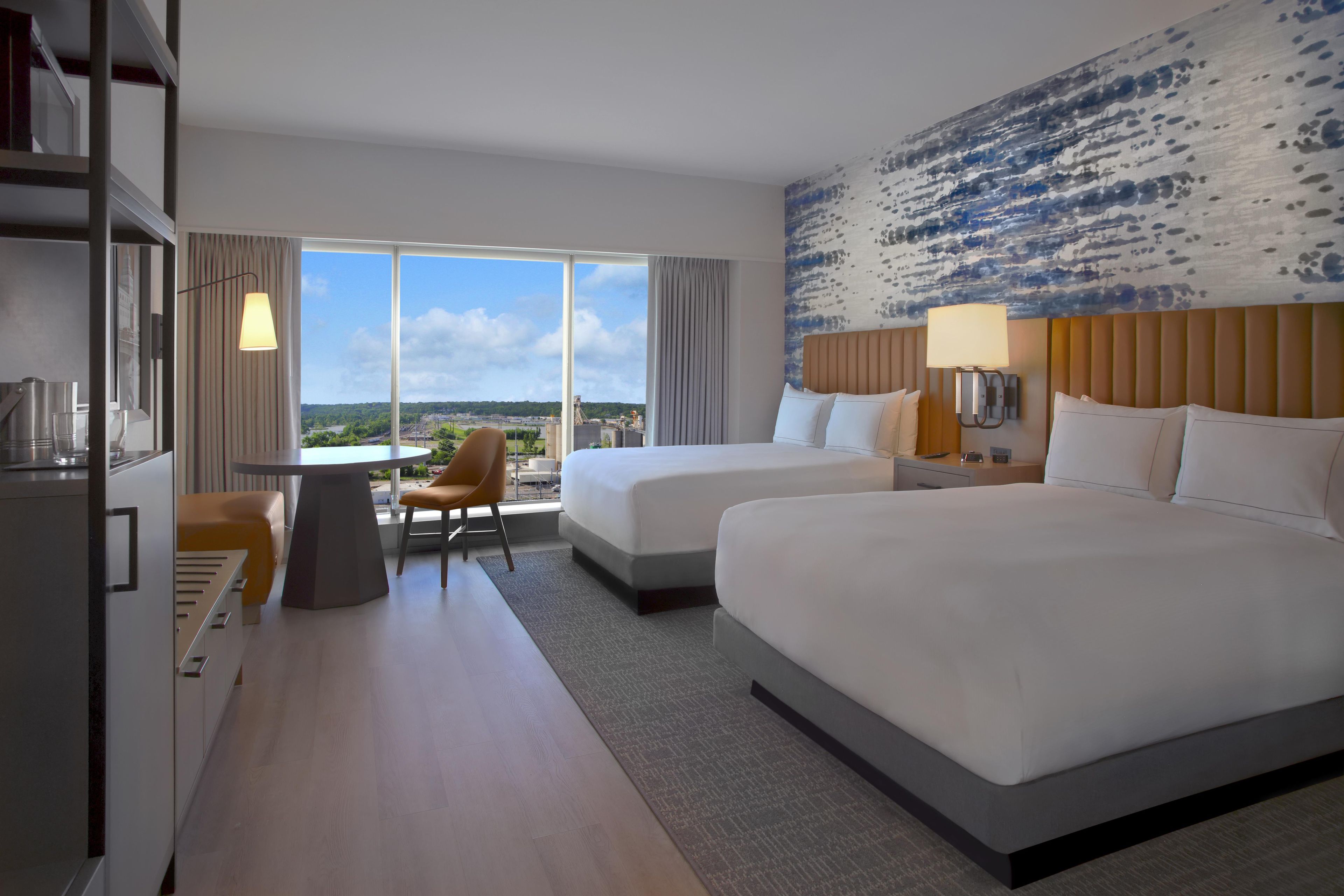 Habitación de hotel con dos camas, un escritorio, una silla y un ventanal con vistas a la ciudad.
