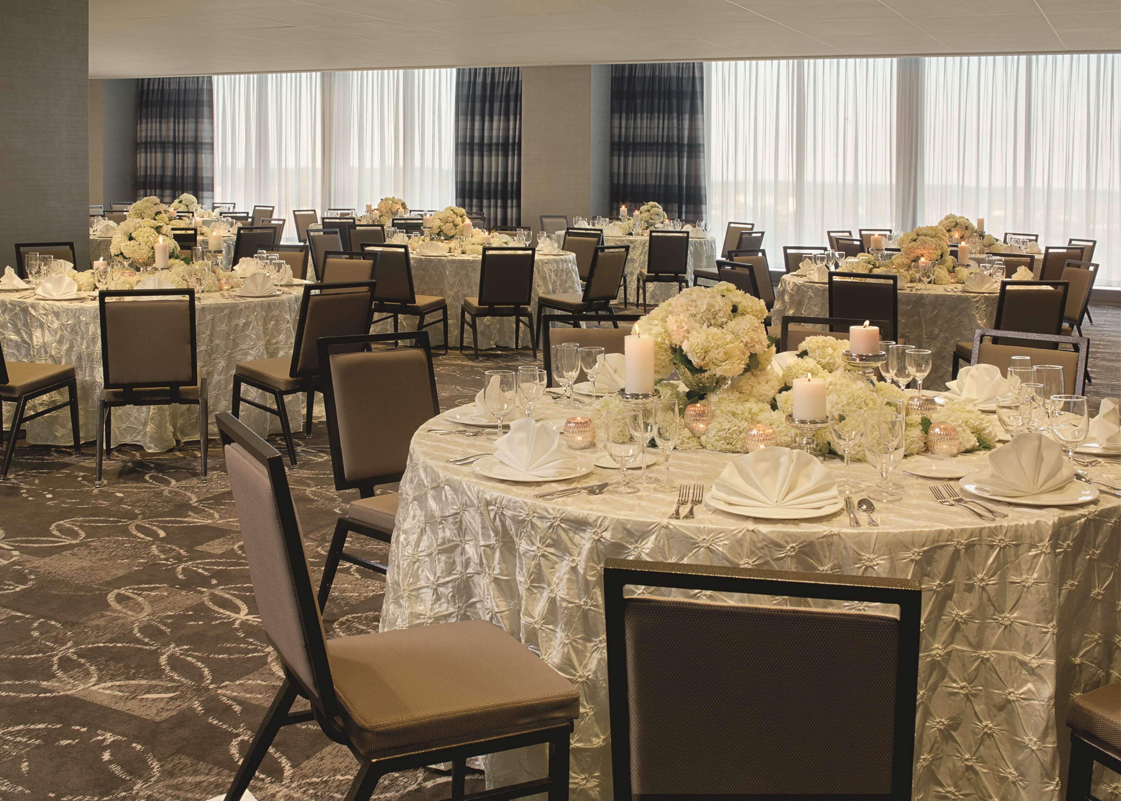 A banquet hall with round tables set for a formal event, featuring white tablecloths, floral centerpieces, and candle lighting.