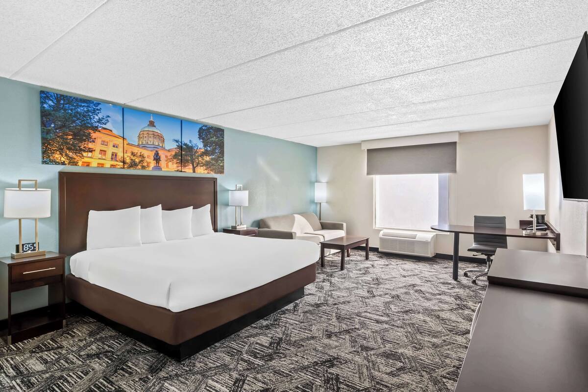 A hotel room with a large bed, a desk, a chair, a sofa, and a painting of a building on the wall.