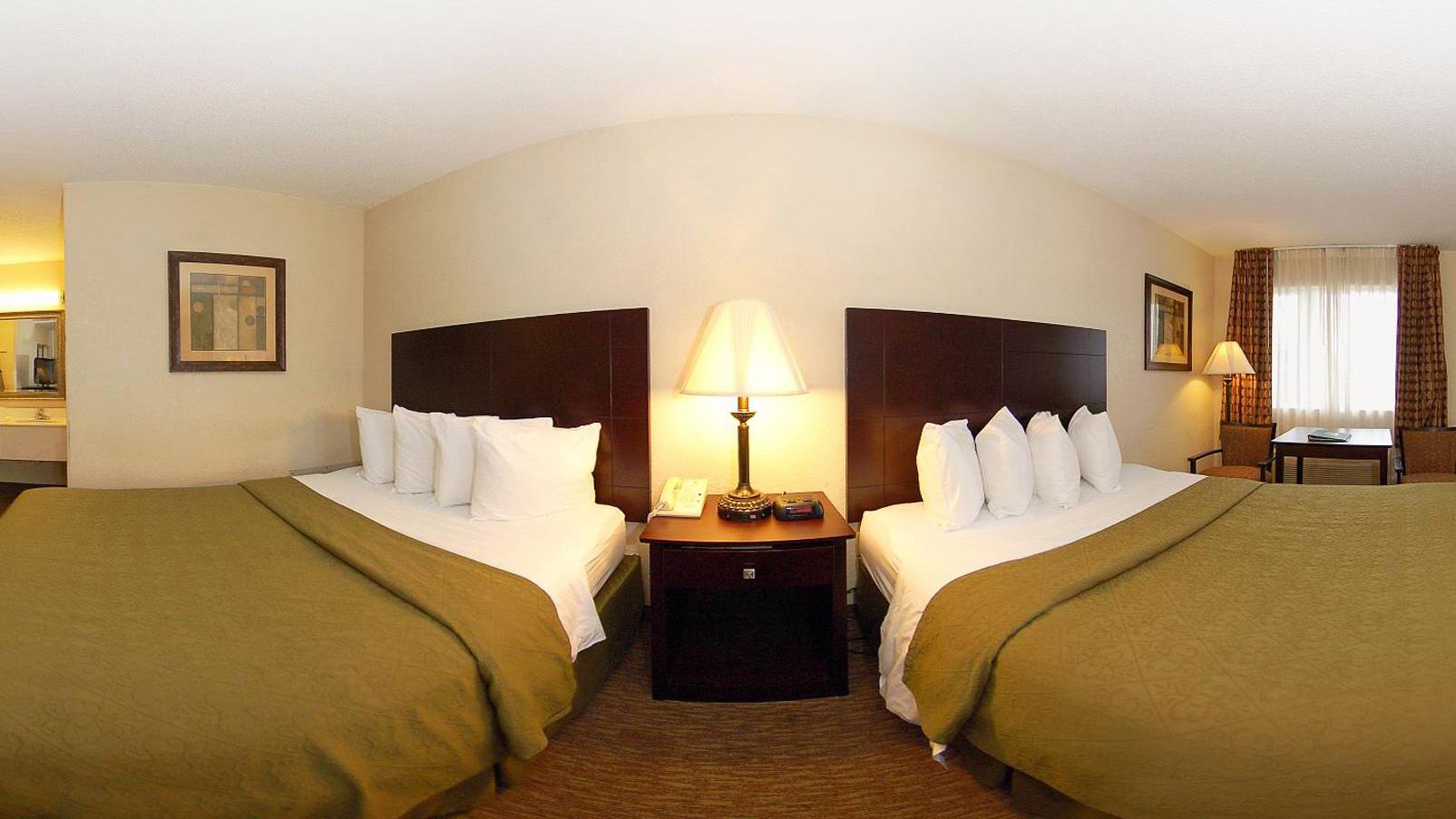 A hotel room with two beds, a nightstand with a lamp, a small table with a chair, and a window with curtains.