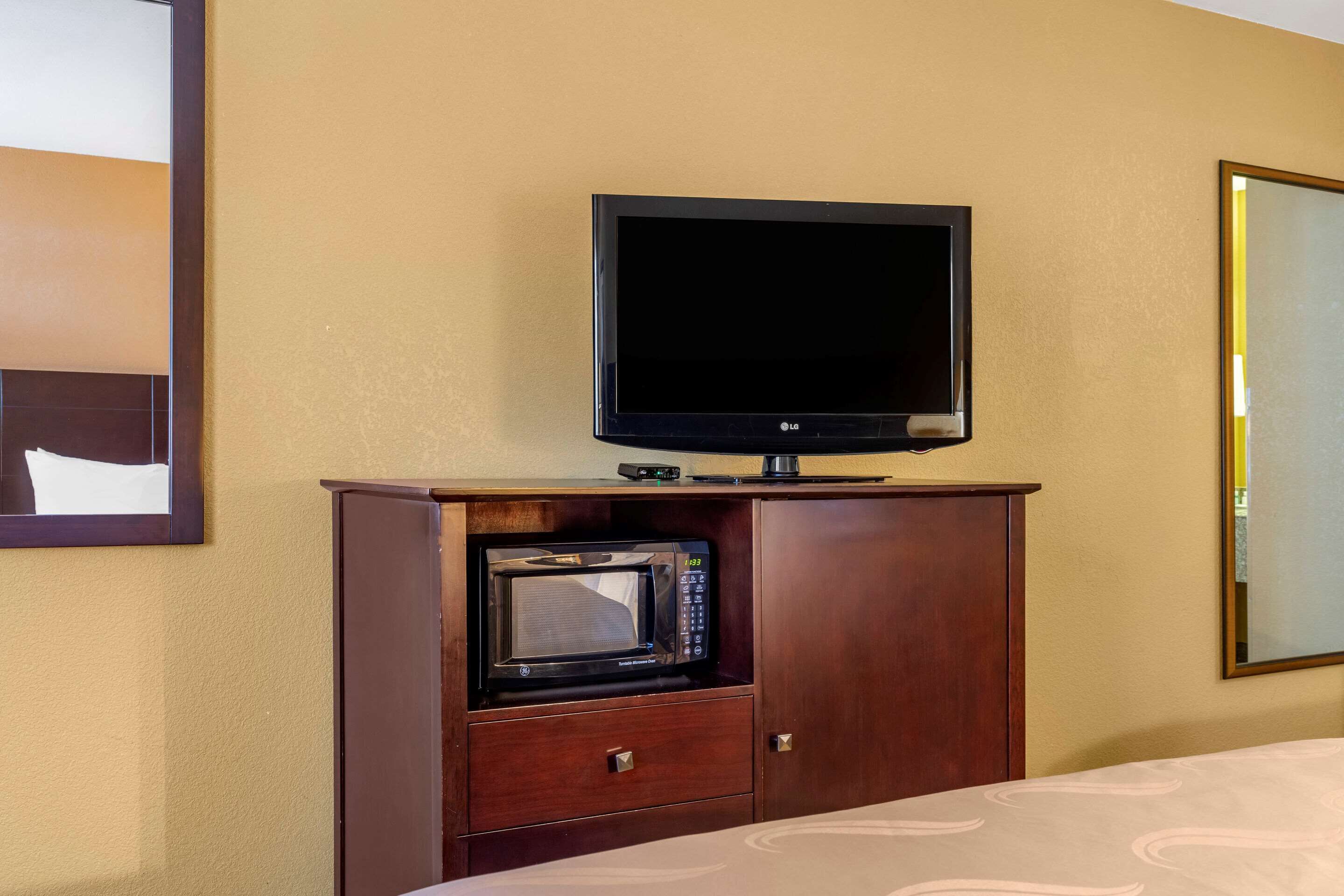 A hotel room with a television mounted above a wooden cabinet, a microwave inside the cabinet, and a bed with a patterned bedspread.