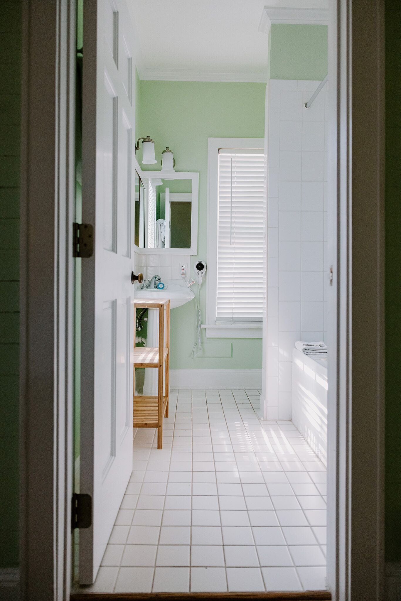 A bathroom with white tiles, a sink, a mirror, and a window with blinds.