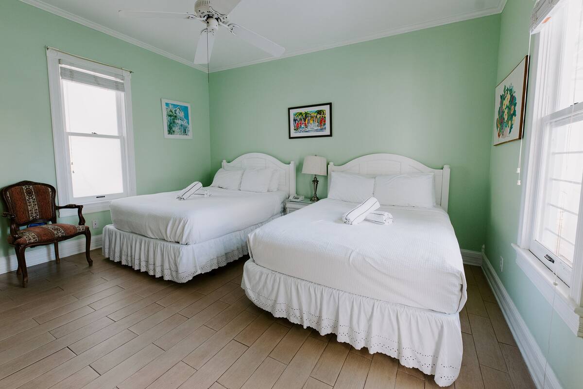 Ein Zimmer mit zwei Betten, einem Sessel, einem Deckenventilator und Kunstwerken an den Wänden.