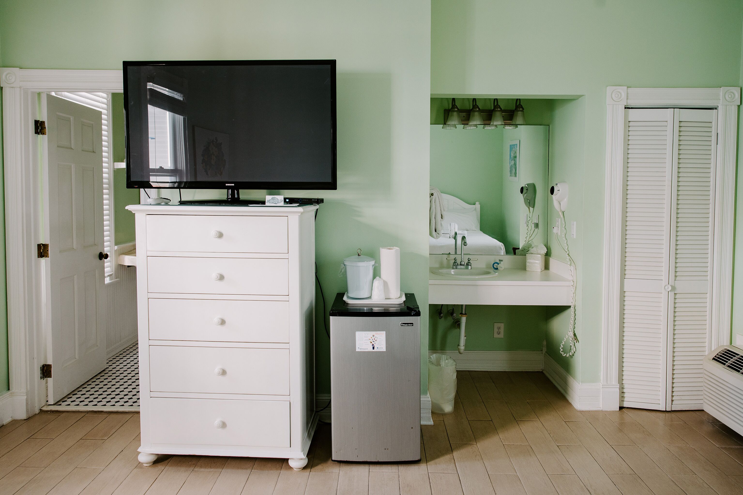 Una habitación con un tocador blanco, un refrigerador pequeño, un televisor encima del tocador, un lavabo y una puerta con una ventana.