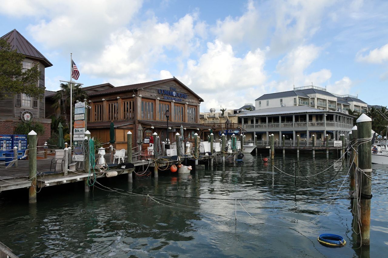 A waterfront restaurant with a dock extending into the water, a flagpole with an American flag, and a building with a sign reading "The Commodore Restaurant."