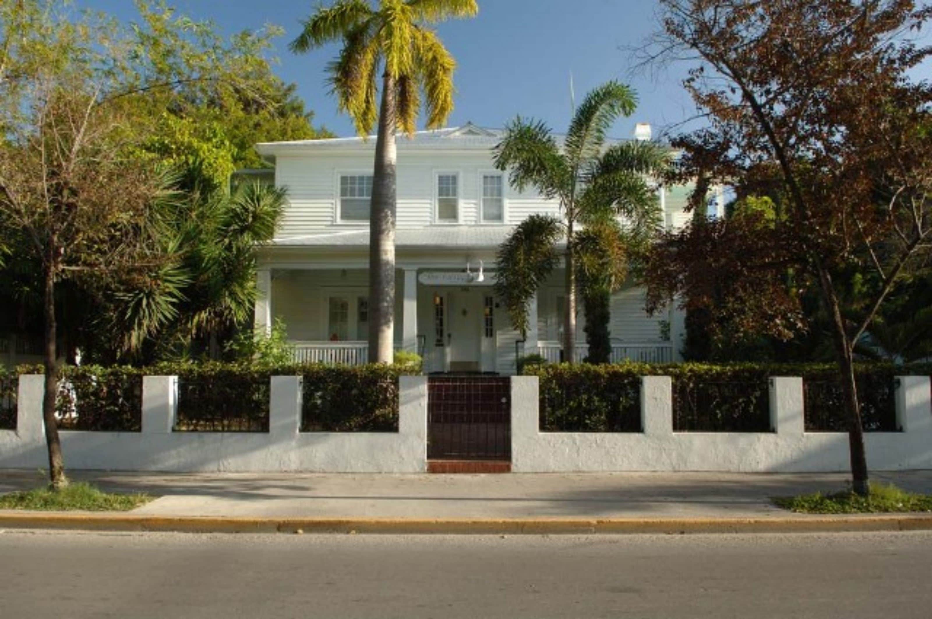 Une maison à deux étages avec un porche à l’avant, entourée d’une clôture blanche et de palmiers.