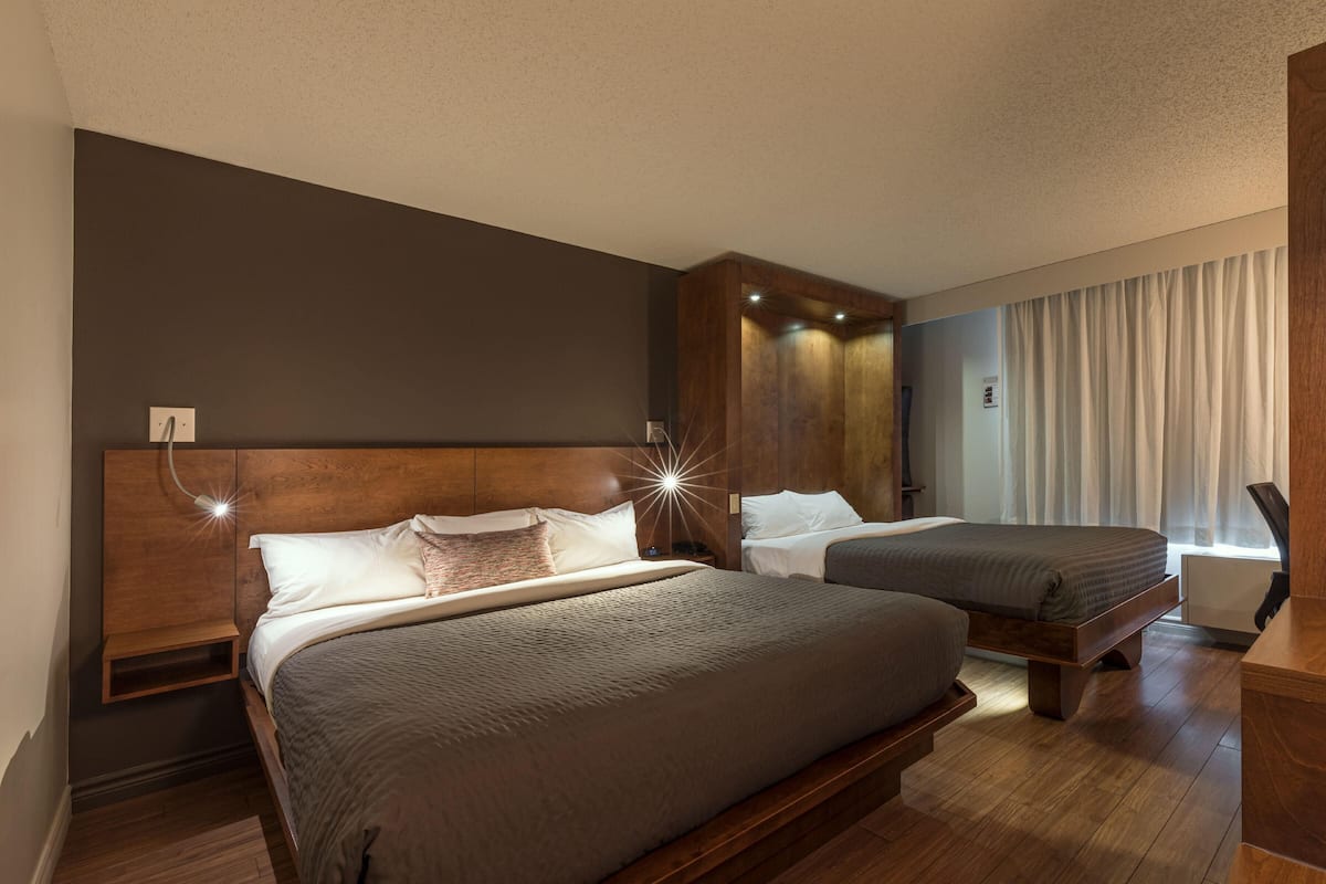 A hotel room with two beds, wooden headboards, a nightstand, a desk, and a large window with curtains.