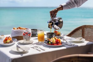 Una mesa de desayuno con diversos platos, una jarra sirviendo zumo de naranja y vista al océano.