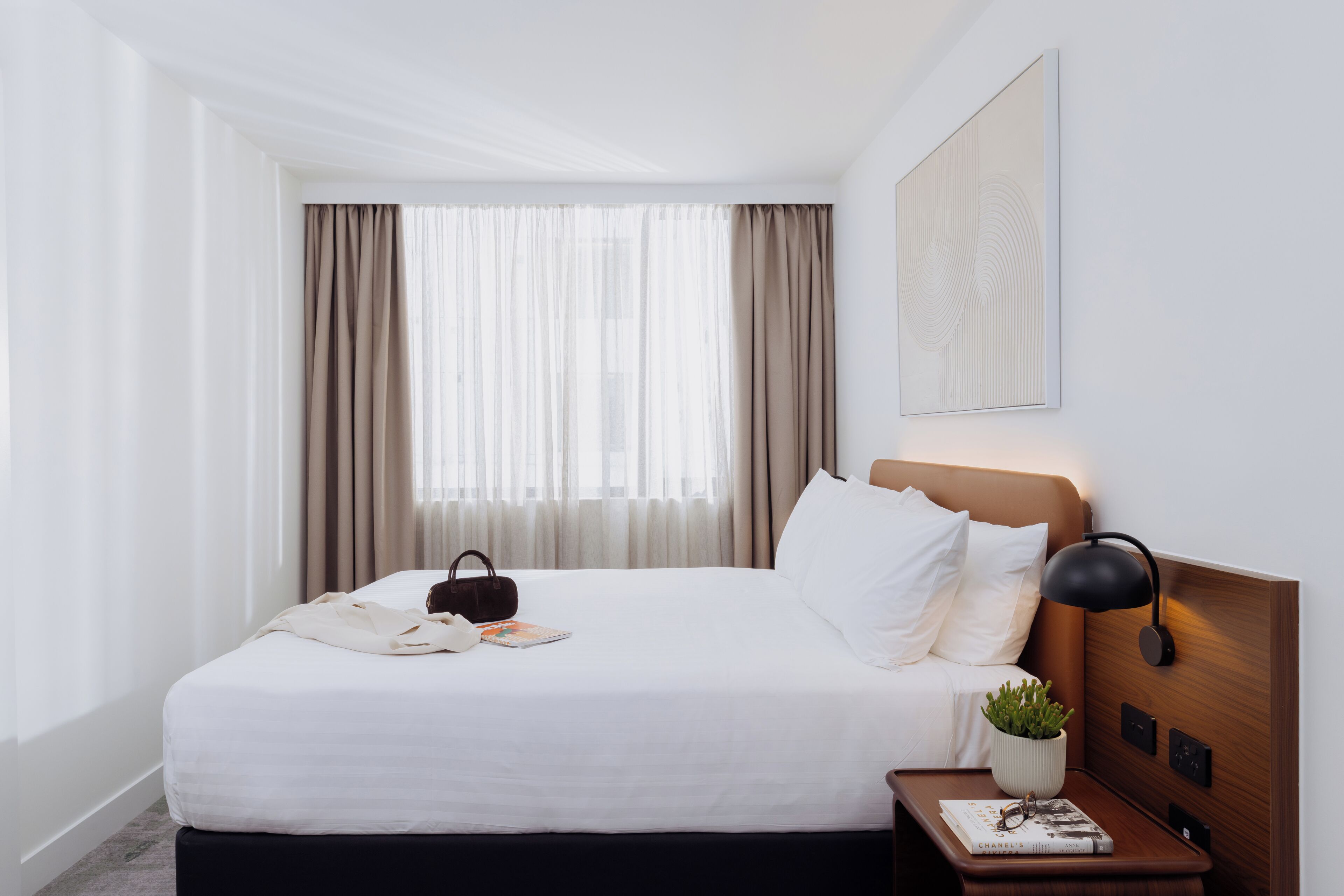 Una habitación de hotel con una cama bien hecha, una mesita de noche con una lámpara, una pequeña planta en maceta y un libro.