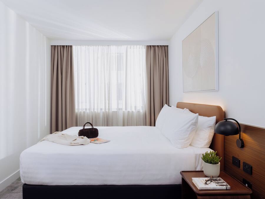 Una habitación de hotel con una cama bien hecha, una mesita de noche con una lámpara, una pequeña planta en maceta y un libro.
