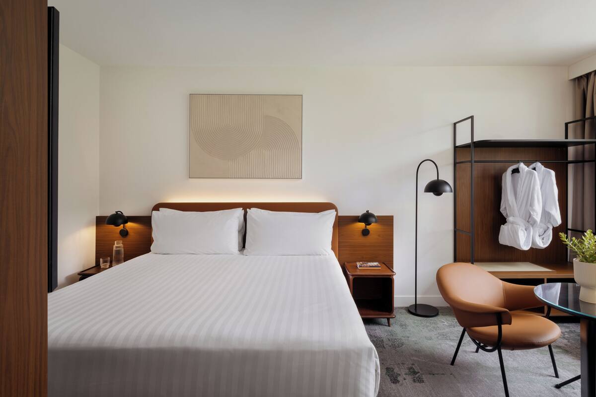 A hotel room with a bed, bedside tables, a chair, a wardrobe, and a wall-mounted artwork.