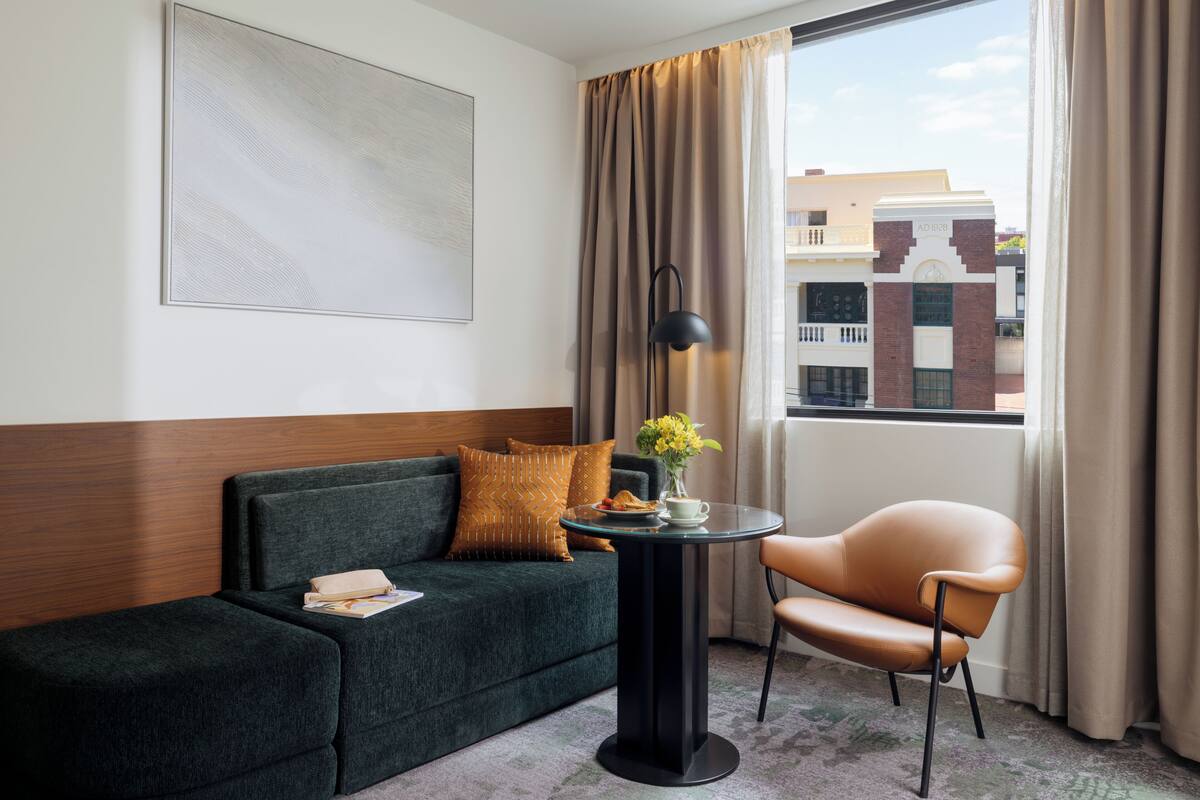 A hotel room with a dark green sofa, a round table with a lamp, a chair, a window with curtains, and a view of buildings outside.