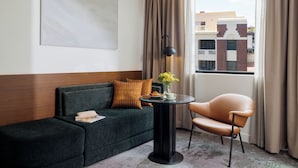 Habitació d'hotel amb un sofà verd fosc, una taula rodona amb una làmpada, una cadira, una finestra amb cortines i vista a edificis exteriors.