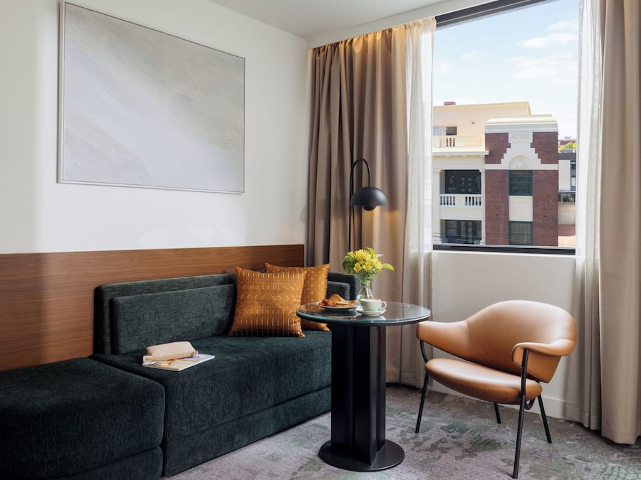 Habitación de hotel con un sofá verde oscuro, una mesa redonda con lámpara, una silla, una ventana con cortinas y vistas a edificios.