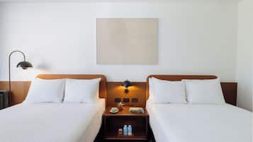 Una habitación de hotel con dos camas, una mesita de noche con lámpara, una pequeña mesa con una taza y un libro, y una obra de arte colgada en la pared.