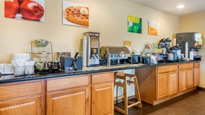 A long counter with various coffee machines and food items, wooden cabinets, and a small stool. - Quality Inn Pueblo North (Pueblo)